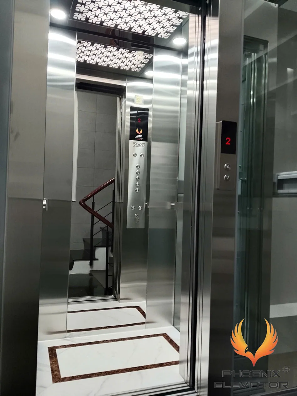 Cabin thang máy quan sát QS07 với vách inox sáng, mảng kính và sàn trắng viền nâu