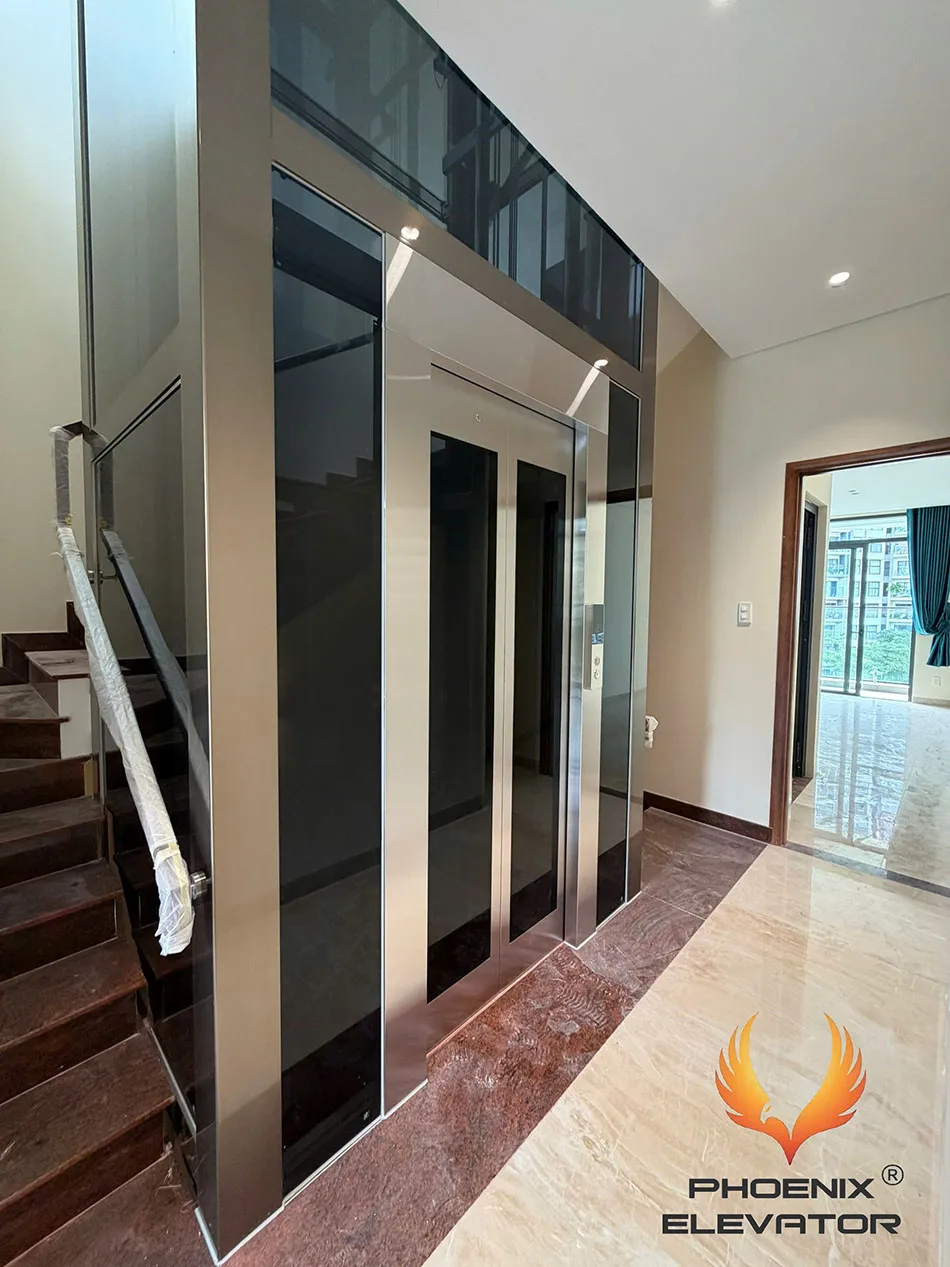 Thang máy khung nhôm khung champagne lắp cạnh cầu thang gỗ trong nhà phố, kính tea và sàn đá marble