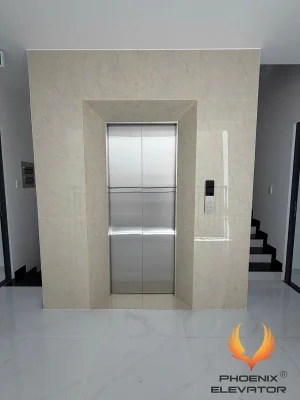 Sảnh tầng thang máy gia đình GD06 ốp đá vân kem tạo khối cổng quanh cửa, bảng gọi tầng mặt kính đen báo tầng