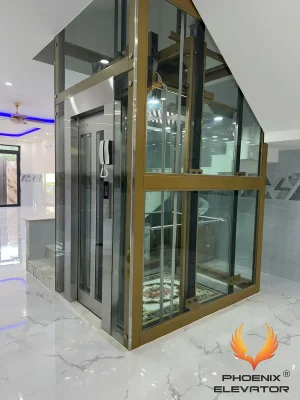 Thang máy quan sát Phụng Hoàng mẫu QS09-2026 với giếng kính cường lực trắng lắp lọt lòng khung nhôm màu vàng đồng, dựng trong nhà phố
